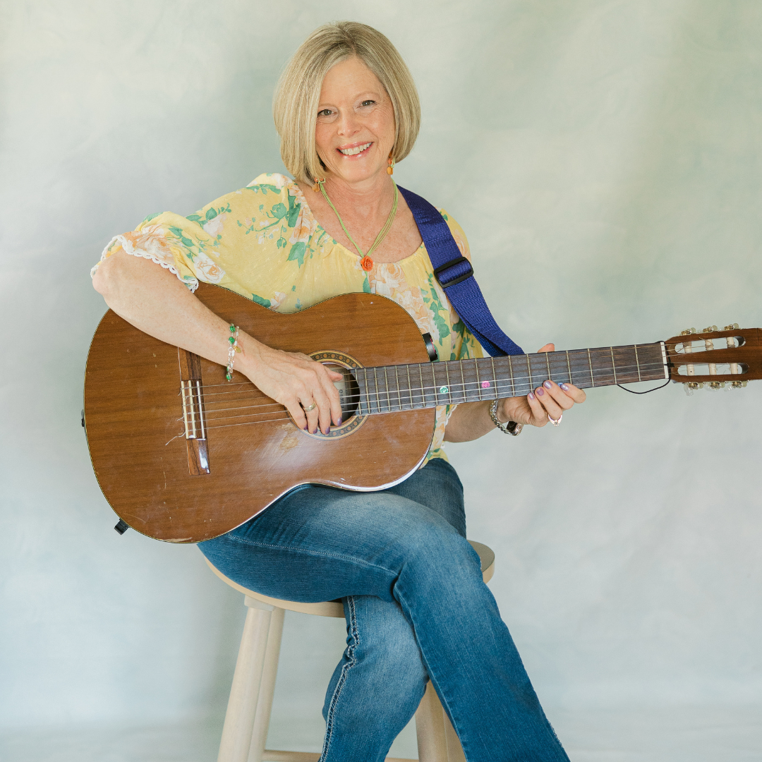 blonde woman in yellow flowery shirt and jeans sitting on a stool holding her guitar and smiling