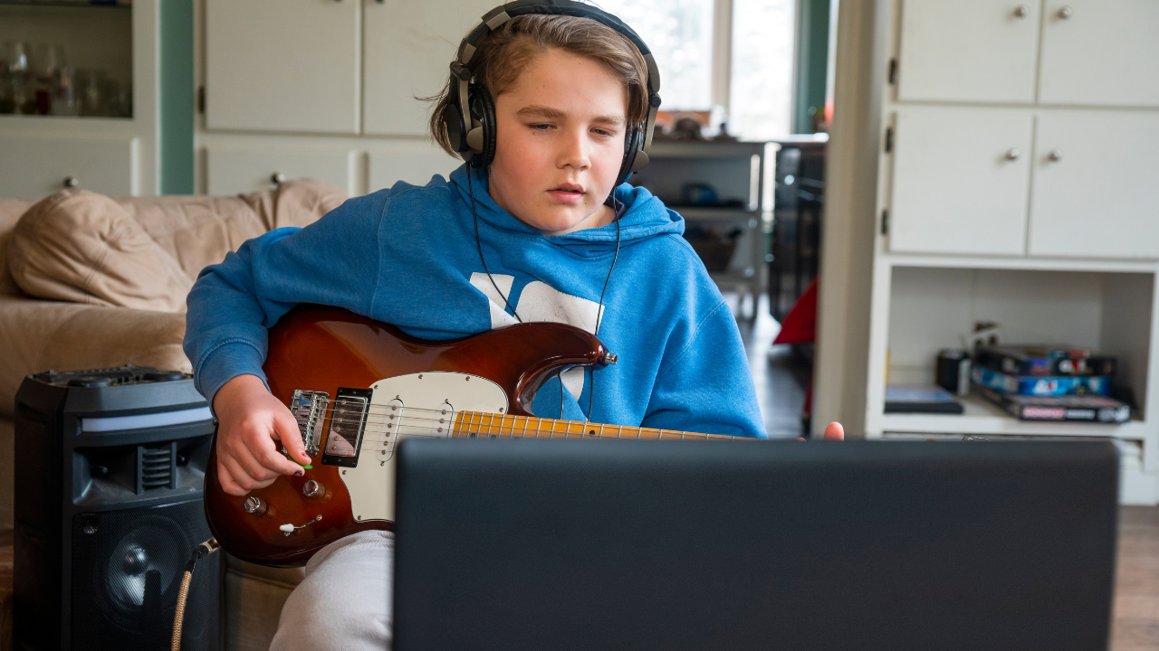 boy with brown hair wearing headphones and holding a guitar taking lessons at the computer with happy kids guitar lessons